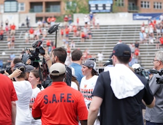 A lively gathering in an outdoor sports venue with a group of people, some wearing matching shirts labeled 'National Champions.' Several individuals are holding cameras and microphones, indicating a media presence. The background shows blurred spectators sitting on bleachers.
