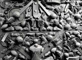 A complex arrangement of various sports equipment and attire, including tennis rackets, soccer balls, basketballs, helmets, shoes, gloves, and a mannequin torso wearing a T-shirt. The items are layered and artistically displayed in a monochrome color scheme, creating an intricate texture and visual pattern.