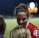 A female athlete in a red sports jersey with the NWSL logo energetically interacts with others on a sports field. The backdrop consists of a darkened night sky and a blurred scoreboard or advertisement display, with soft, colorful lights.