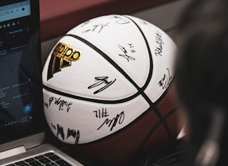 A signed basketball rests against a laptop on a table. A person's hand is visible, possibly using the laptop, and a microphone can be seen close by, suggesting an interview or commentary setting.
