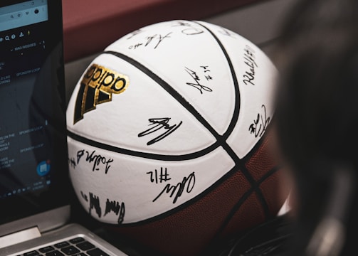 A signed basketball rests against a laptop on a table. A person's hand is visible, possibly using the laptop, and a microphone can be seen close by, suggesting an interview or commentary setting.