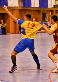 A dynamic indoor sports scene with players in motion on a court. Two athletes in blue and yellow uniforms are engaged in an intense handball game, with one player gripping the other's jersey. The background shows other players, several seated people, and advertisements on the walls.