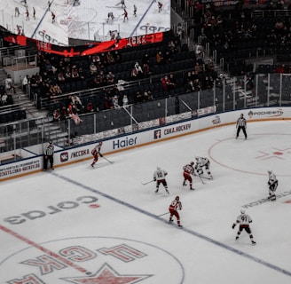 A professional ice hockey game is in progress inside an arena, with players in red and white jerseys actively engaging on the rink. The scoreboard above displays the score and game details, while spectators are seated in the stands. A large screen shows a live feed of the match action.