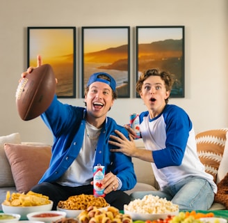 Two young men sitting on a sofa look excited as they engage in watching something, possibly a sports game. One of them, wearing a blue cap and jacket, holds an American football and a can of soda. The other, wearing a blue and white baseball shirt, appears surprised and is also holding a can. In front of them, there is a table covered with various snacks, including chips, salsa, pretzels, popcorn, and a vegetable platter. Three framed photographs of a landscape hang on the wall in the background.