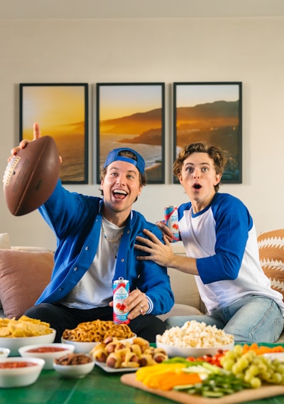 Two young men sitting on a sofa look excited as they engage in watching something, possibly a sports game. One of them, wearing a blue cap and jacket, holds an American football and a can of soda. The other, wearing a blue and white baseball shirt, appears surprised and is also holding a can. In front of them, there is a table covered with various snacks, including chips, salsa, pretzels, popcorn, and a vegetable platter. Three framed photographs of a landscape hang on the wall in the background.