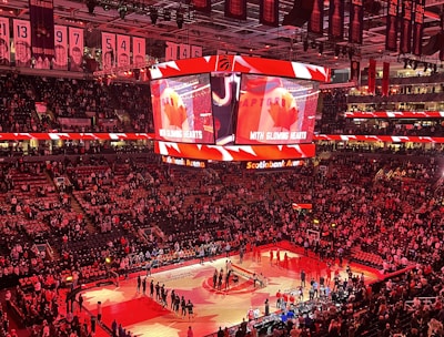 An indoor sports arena filled with spectators and a basketball court illuminated by red lighting. The scoreboards and video screens display promotional content, with numerous banners and championship flags hanging from the ceiling. Players and officials are gathered on the court, possibly in preparation for a game or event.