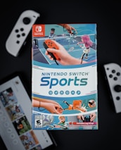 A box of Nintendo Switch Sports is prominently featured, surrounded by various Nintendo Switch controllers and a screen displaying a related interface. The box art depicts animated characters playing sports, such as tennis, with vibrant colors and action-oriented poses.
