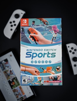 A box of Nintendo Switch Sports is prominently featured, surrounded by various Nintendo Switch controllers and a screen displaying a related interface. The box art depicts animated characters playing sports, such as tennis, with vibrant colors and action-oriented poses.