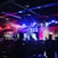 A dimly lit indoor arena filled with an audience. Bright stage lights in red and blue illuminate multiple large screens displaying graphics.