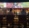 A group of people sit at a bar with their backs to the camera, watching multiple sports games displayed on large screens. The bar counter is lined with various bottles of liquor, and the atmosphere suggests a casual and relaxed social setting.