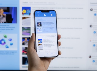 A hand holding a smartphone displaying a news website, with a blurred large computer screen in the background also showing similar content. The webpage on the phone features articles with images and text.