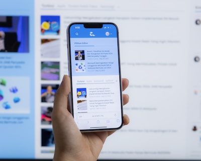 A hand holding a smartphone displaying a news website, with a blurred large computer screen in the background also showing similar content. The webpage on the phone features articles with images and text.