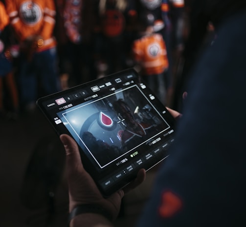 A person is holding a tablet that displays a video or image of a red and blue emblem on the screen. In the background, several people wearing dark and orange colored clothing, possibly sports jerseys, are partially visible. The scene is dimly lit, suggesting an indoor or evening setting.