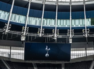 A modern stadium interior featuring a large digital screen with a logo prominently displayed. The seating areas are empty, with dark-colored seats and some structural elements like beams and glass walls visible. A combination of dark blue and neutral tones are prominent.