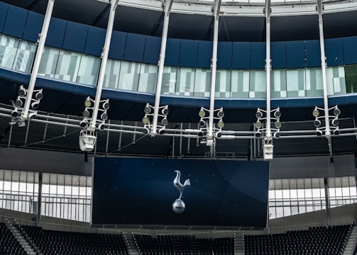 A modern stadium interior featuring a large digital screen with a logo prominently displayed. The seating areas are empty, with dark-colored seats and some structural elements like beams and glass walls visible. A combination of dark blue and neutral tones are prominent.